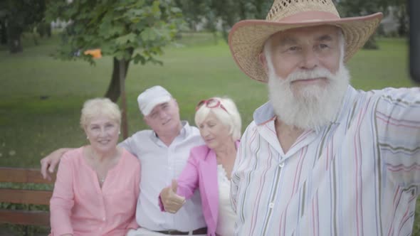 Adult Man in a Hat and Gray Beard Makes a Selfie Against the Background of His Friends Sitting