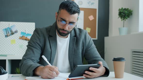 Serious Entrepreneur Writing and Looking at Tablet Screen Busy with Work in Office