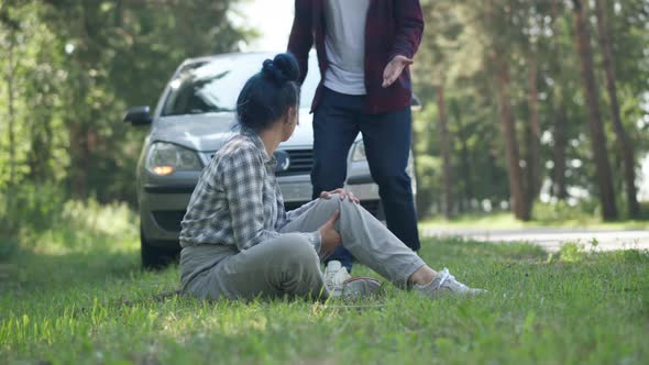 Angry Knocked Down Woman Sitting on Ground Rubbing Knee with Man Running From Car at Background