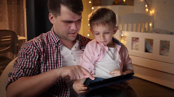 Portrait of Little Boy Using Digital Tablet Computer with His Young Father at Night Before Going To