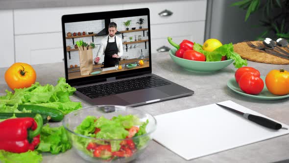 Laptop with Man Chef in Screen Stands on Kitchen Table Near Food Vegetables