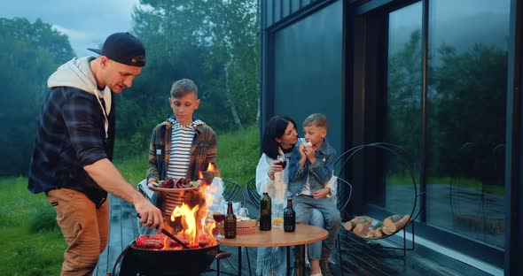 Family with Children Having Family Picnic Uutdoors in the Yard, Dad Preparing Sausages on Grill 