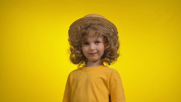 A Cute Little Curly Girl in a Straw Hat is Smiling Thoughtfully in Yellow Studio