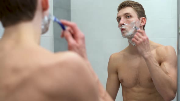 Young Caucasian Man Look in Bathroom Mirror Apply Foam Gel Shaving with Razor After Shower