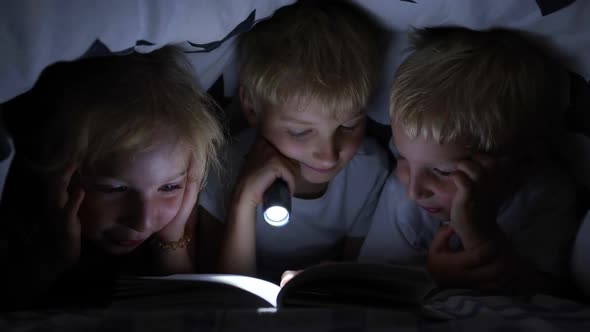 Three Young Children Are Reading a Book with a Flashlight Under the Covers at Night.