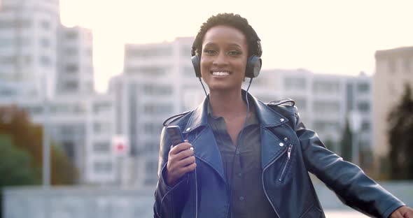 Happy African American Teenage Girl Wears Leather Jacket and Headphones, Holds Smartphone in Hands