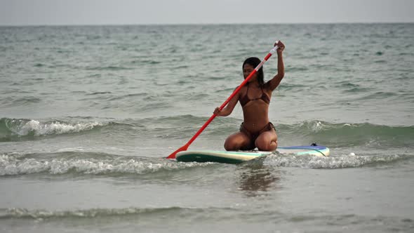 Young Woman In Bikini Kneeling On Paddleboard And Rowing In Sea
