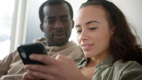 Young Smiling Couple Relaxing At Home On Sofa Checking Social Media On Mobile Phones Together