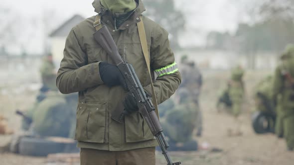 Unrecognizable Male Soldier Standing Outdoors with Armed Rifled Gun As Blurred Recruits Resting at