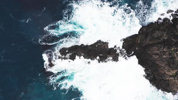 Aerial View of Waves Break on Rocks of Faroe Islands Cliffs in a Blue Ocean