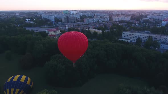 Red Hot Air Balloon Floating Over City Green Area at Dawn, Festival Early Flight