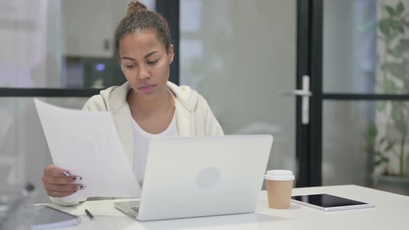 African Woman with Laptop Reading Documents in Office