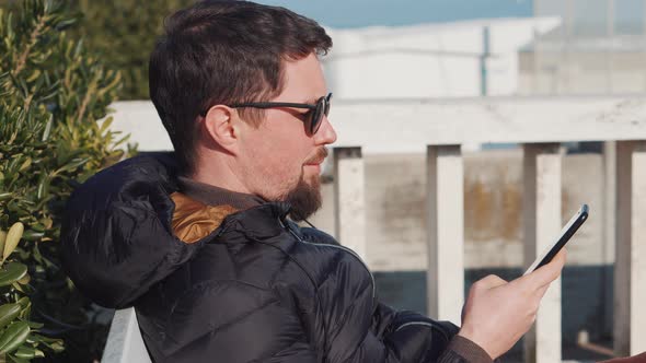 Townsman Looks Through News on Smartphone Sitting on Bench