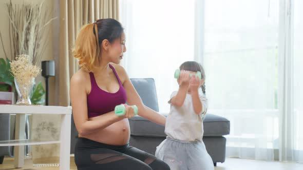 Asian young pregnant woman use dumbbells workout with little daughter.