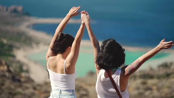 Silhouette of Two Young Girls Standing on Cliff with the View on Blue Sea Lagoon Holding Their Hands