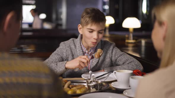 Portrait of Happy Boy Enjoying Dinner with Parents Sitting in Restaurant Chewing Sausage Smiling