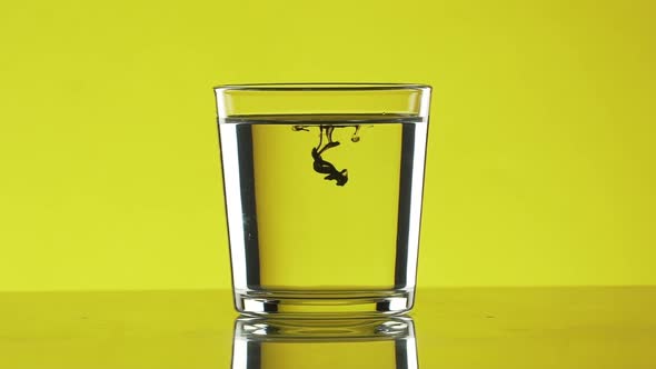 Two Small Drops of Black Ink Dripping Into Clear Water in Glass Standing on Yellow Background in