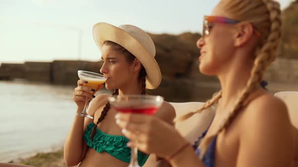 Two Beautiful Young Girls Relaxing Talking Laughing Drinking Cocktails at the Seaside