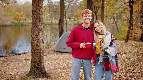 Young Loving Man and Woman Smiling and Hugging Holding Cups of Tea While Standing in Autumn Forest