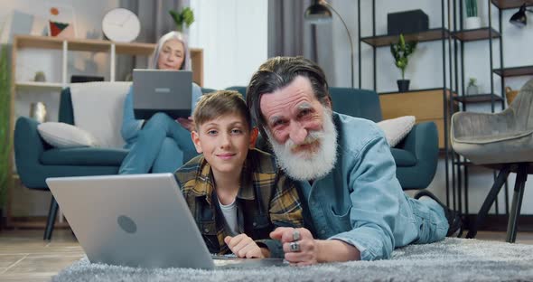 Senior Grandfather Lying on the Floor Together with His 12-Aged Grandson and Using Laptop
