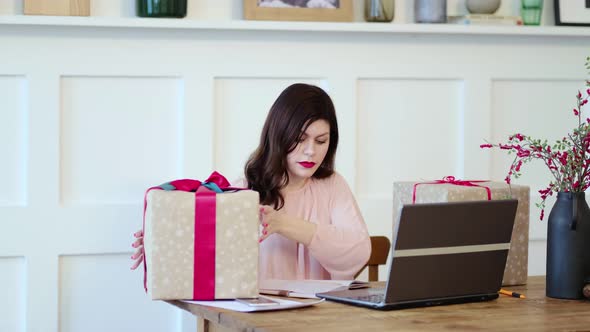 Pretty Young Lady Sitting at Desk and Typing on Laptop and Talking with Present Boxes Icons