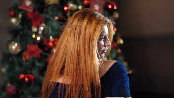 Beautiful Red-haired Woman Looks at Camera Against the Background Christmas Tree