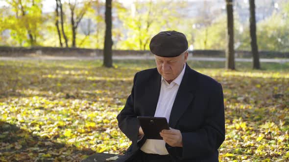 Joyful Senior Man Resting on a Park Bench Typing on the Tablet and Telling