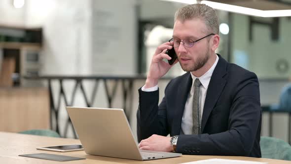 Hardworking Businessman with Laptop Talking on Smartphone in Office 