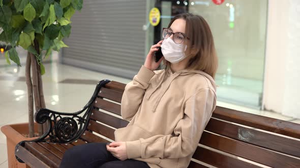 A Young Woman in a Medical Mask Sits on a Bench in a Shopping Center and Talks on the Phone. The