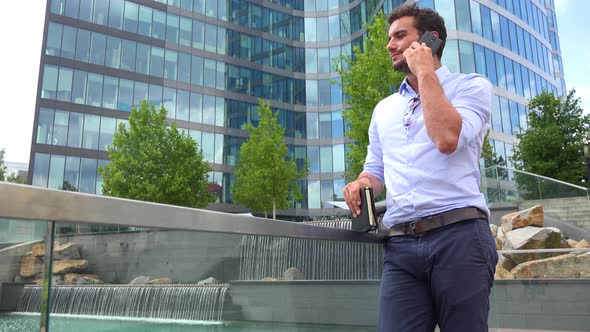 A Young Man Talks on a Smartphone in an Urban Area, Artificial Waterfalls and an Office Building