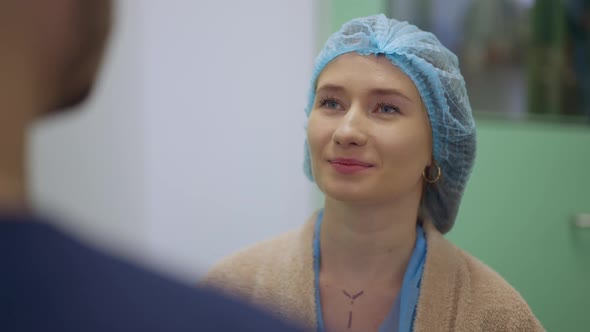 Portrait of Confident Young Caucasian Beautiful Woman in Surgical Cap Talking with Unrecognizable