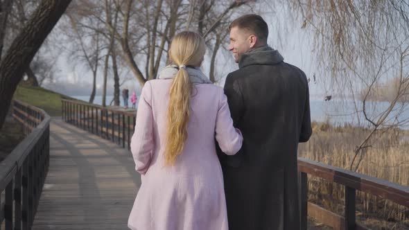 Back View of Adult Caucasian Man and Woman in Elegant Coats Walking in Park. Positive Married Couple