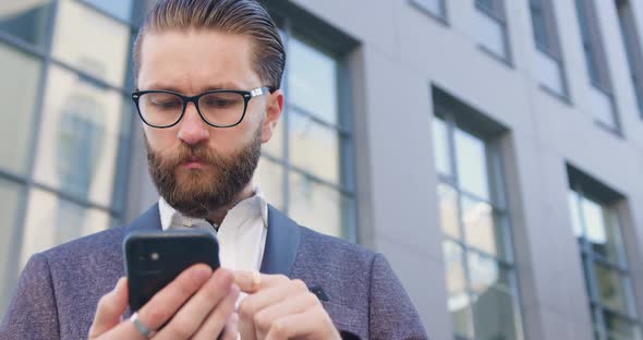 Office Manager Standing Near Modern Office Building Scrolling and Reading News on His Smartphone