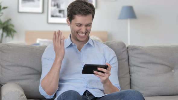 Young Man Cheering Success on Smartphone while Sitting on Couch