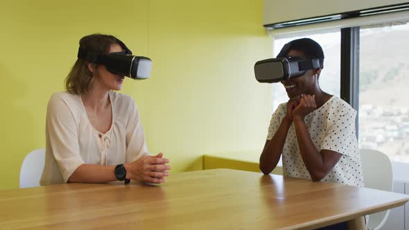 Two diverse female coworkers sitting at desk, testing vr googles