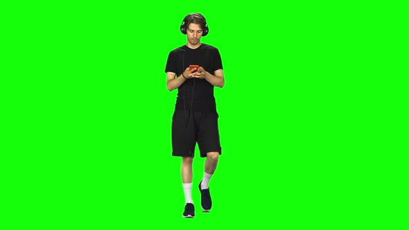 Brunette Man in Big Headphones Goes and Texting with Smartphone on Green Screen at Studio.