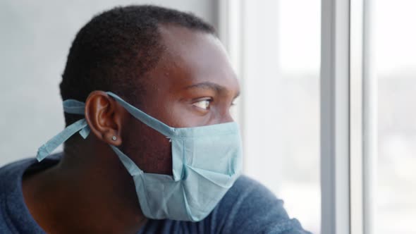 a Man with a Protective Mask on His Face During Quarantine Sits By the Window with a Smartphone