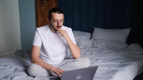 Serious Bearded Man in Glasses and a Tshirt is a Freelancer Sitting on a Bed with a Laptop in an