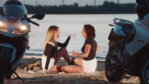 Two Women Friends Motorcycle Riders Sitting on the Beach and Talking at Late Sunset Evening