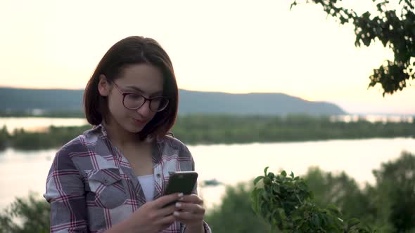A Young Woman Stands on a Hill Against the Background of the River and Mountains with a Phone in Her