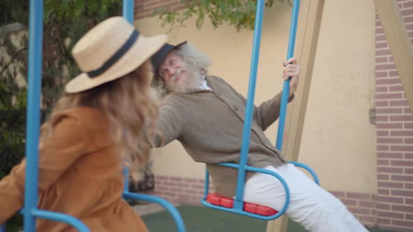 Side View of Happy Elegant Grandfather Enjoying Weekend with Granddaughter Having Fun on Swings