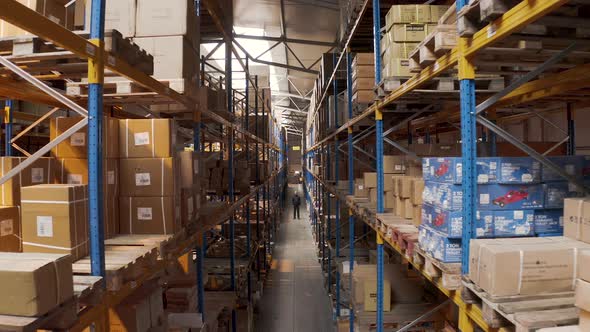 Man standing in a long storage warehouse aisle with boxes,aerial zoom.