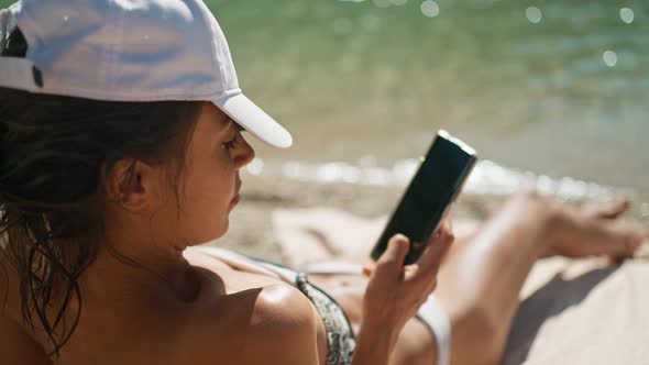 Back View Relaxed Woman Tourist in Bikini Sunbathes on Sea Beach Close to Water Unlocks Her Mobile