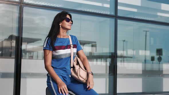 Travel Woman Sitting on Suitcase Looking for Watch Waiting Departure at Airport Terminal Exterior