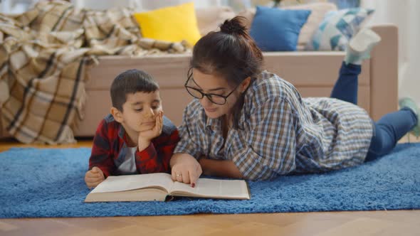 Little Boy with Mother Lying on Carpet and Reading Book in Living Room