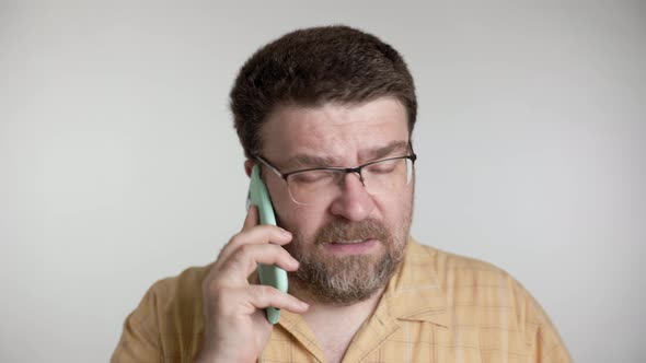 Man with a beard and glasses is angry and aggressively talking on a smartphone on a gray background.
