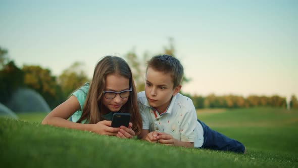 Children Using Smartphone in Park. Siblings Lying on Green Grass in Meadow