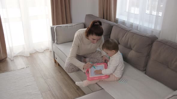 Caring Young Mother or Nanny Helping Her Cute Daughter Teach the Baby Draw a Picture with Pencils