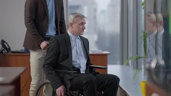 Side View Portrait of Senior Caucasian Disabled Man on Wheelchair Talking with Unrecognizable Male