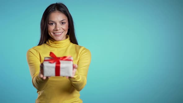 Mixed race woman gives gift and hands it to the camera. She is happy, smiling. Girl on blue